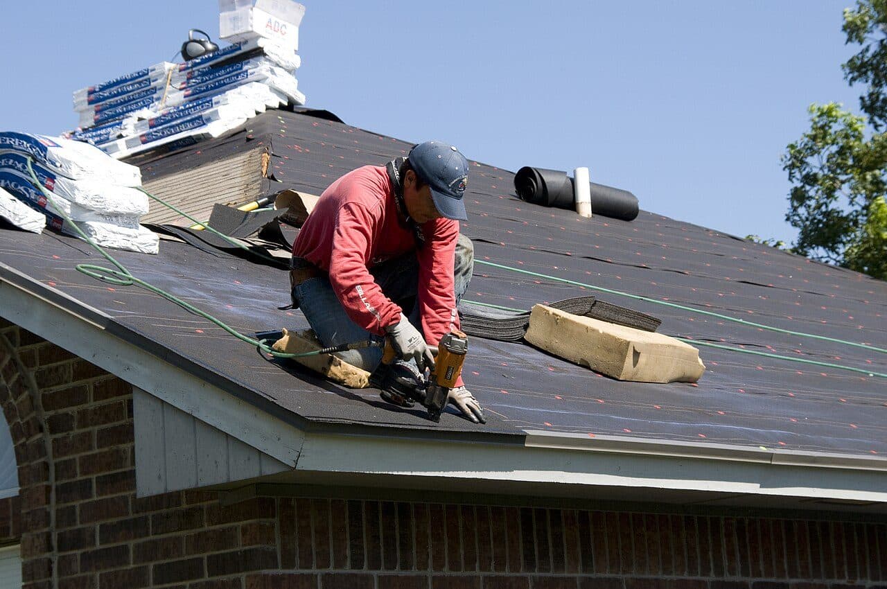 Roofer in a red shirt nailing architectural shingles on a brick home